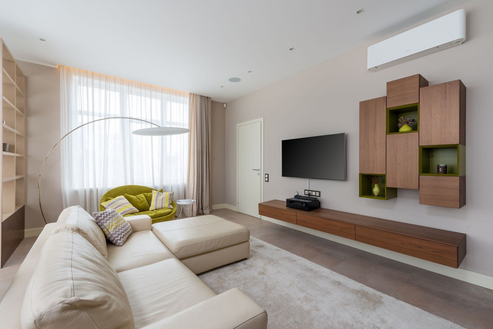 Woonkamer met een crèmekleurige hoekzetel, een tv aan de muur en een witte binnenunit van een airco hoog boven houten hangkastjes