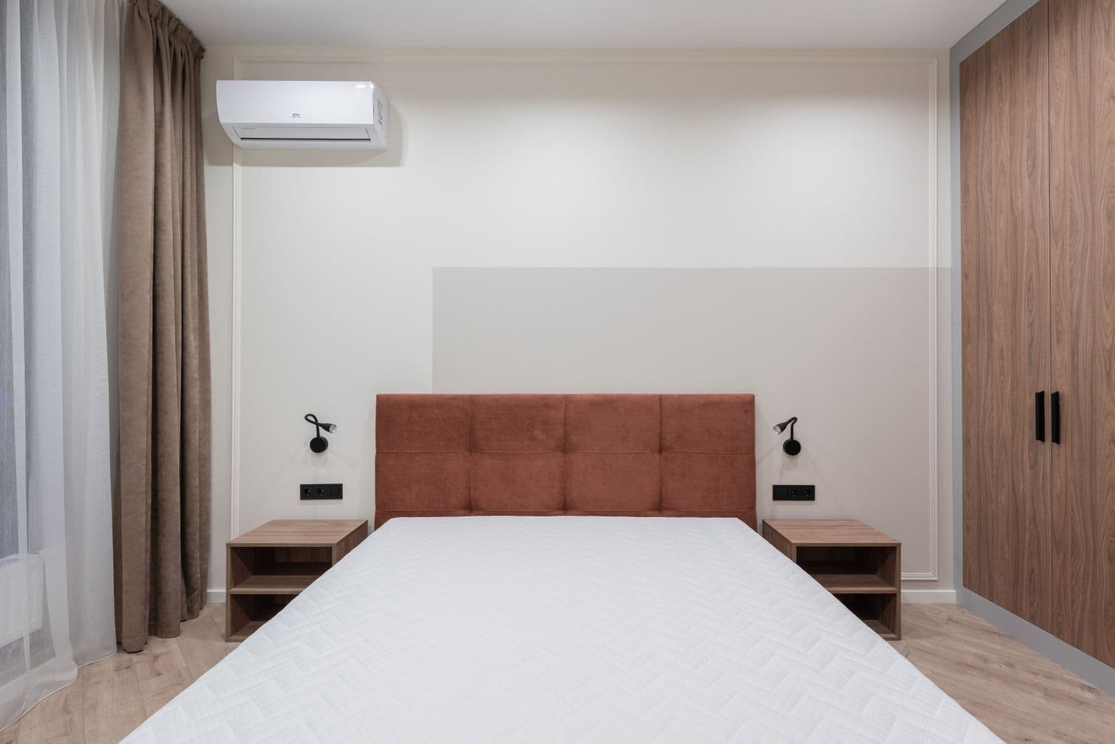 Slaapkamer met een tweepersoonsbed met bruin hoofdbord en een witte binnenunit van een airco hoog aan de muur, naast een bruin gordijn